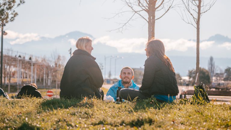 Trois personnes assises sur l'Esplanade de l'EPFL: deux filles de dos, et garçon en tailleur de face. L'Agora Lombard Odier et les Alpes en arrière-plan © Adrien Buttier / EPFL 2024
