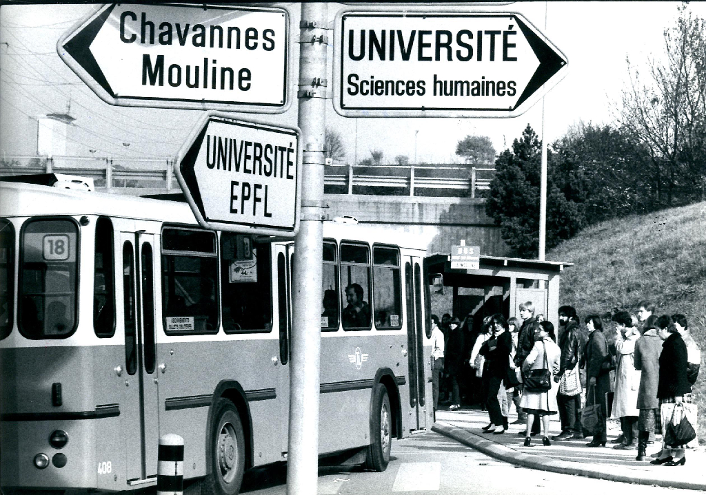 Au premier plan se trouve des panneaux de circulation routière indiquant les directions suivantes : « Université EPFL », « Université Sciences Humaines » et « Chavannes Mouline ». En arrière-plan, les étudiants à droite de l’image attendent le bus qui arrive à gauche de l’image.