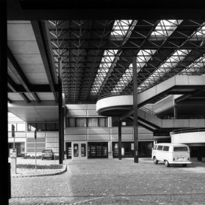 Photographie en noir et blanc de la toiture métallique en treillis tridimensionnels de l’école. Cette structure est portée par des poteaux en métal. La photo est prise au niveau de la rue. On voit sur la droite en arrière-plan les escaliers.