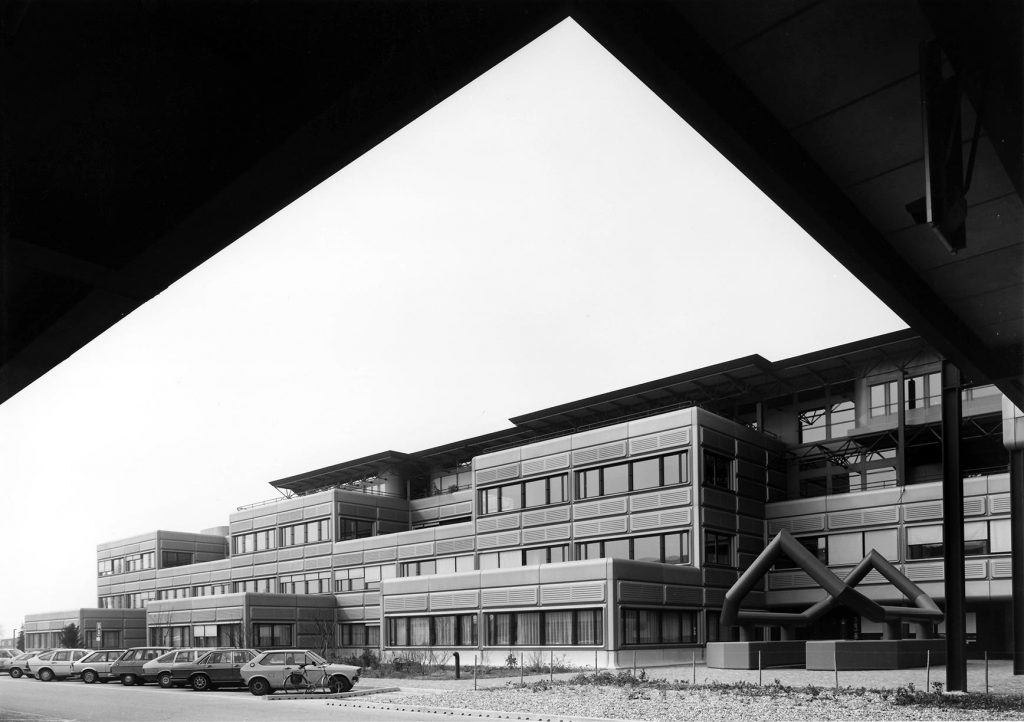 Photographie en noir et blanc prise sous la Méridienne à l’EPFL. Les façades sont revêtues de caissons métalliques arrondis aux angles.