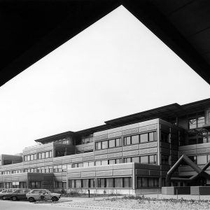 Photographie en noir et blanc prise sous la Méridienne à l’EPFL. Les façades sont revêtues de caissons métalliques arrondis aux angles.