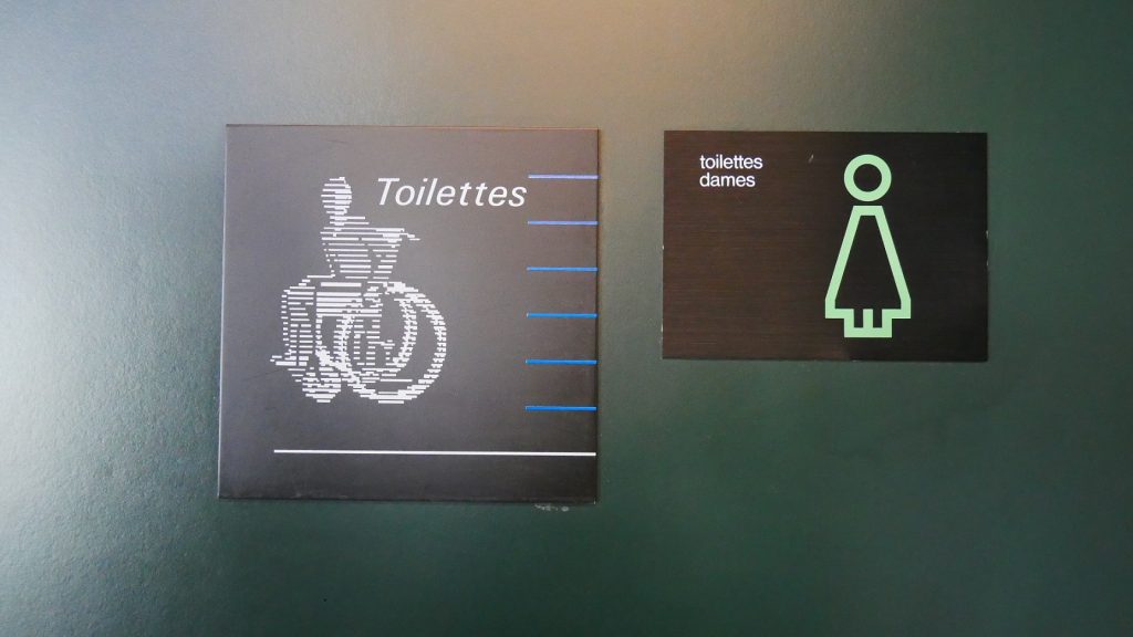 Photographie de deux panneaux différents pour indiquer les toilettes en 2019. Côte-à-côte, l’un date de l’époque Calame et l’autre de la période Ballmer.