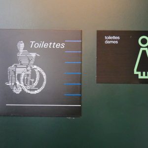 Photographie de deux panneaux différents pour indiquer les toilettes en 2019. Côte-à-côte, l’un date de l’époque Calame et l’autre de la période Ballmer.