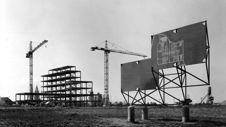 Vue du chantier de l’EPFL en avril 1975 montrant la structure des halles de chimie en construction, deux grues et deux panneaux de chantier au premier plan.