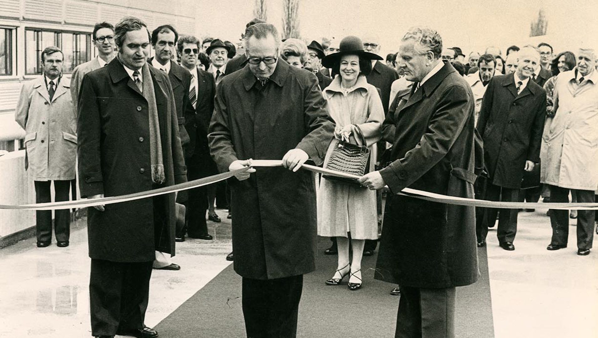 Un homme au centre de l’image coupe un ruban d’inauguration. Il est entouré de deux autres hommes se tenant sur le côté et d’une foule de personnes se tenant derrière lui.
