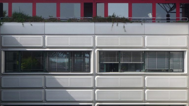 Vue d'une partie de la façade montrant au niveau inférieur quatre fenêtres disposées de manière symétrique entrecoupées verticalement de panneaux métalliques gris ; et au niveau supérieur la terrasse couverte d'un treillis bleu triangulaire et une façade rouge en retrait.