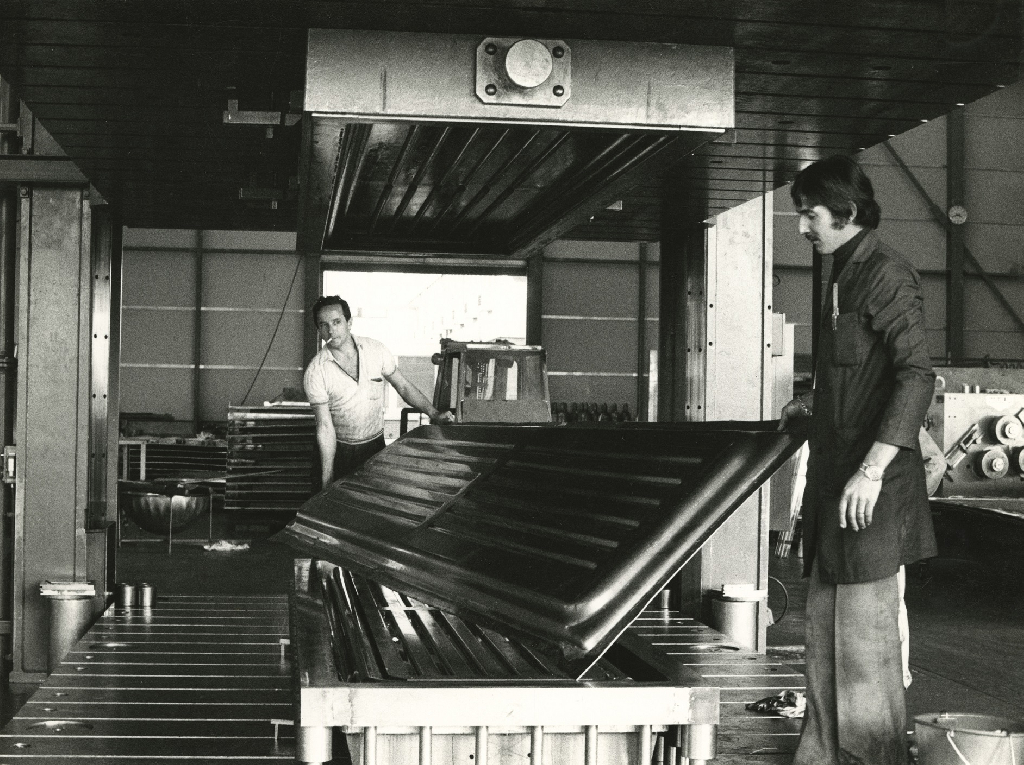 Vue sur un atelier dans lequel deux hommes soulèvent un panneau métallique hors d'une presse à emboutir, face à l'appareil photographique. Photographie en noir et blanc.