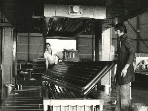 Vue sur un atelier dans lequel deux hommes soulèvent un panneau métallique hors d'une presse à emboutir, face à l'appareil photographique. Photographie en noir et blanc.