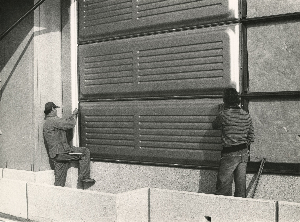 Deux hommes de dos se tenant à chaque extrémité d'un panneau métallique le fixent en bas d'une façade en construction, sous une série d'autres panneaux déjà installés.