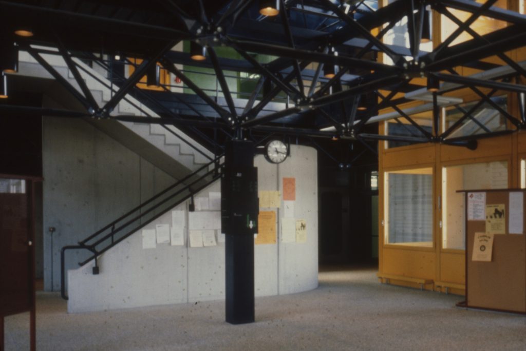 La photographie montre un hall d'entrée de l'école avec au centre un pilier soutenant le treillis bleu en acier, et une cage d'escalier en béton.