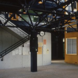 La photographie montre un hall d'entrée de l'école avec au centre un pilier soutenant le treillis bleu en acier, et une cage d'escalier en béton.