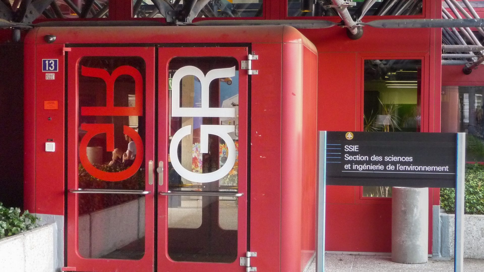 Entrée du bâtiment GR avec textes de signalisation sur les portes et sur un panneau indépendant.