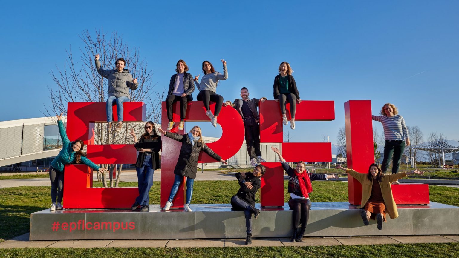 12 LCE staff members standing on a large red EPFL statue outdoors, posing in silly poses