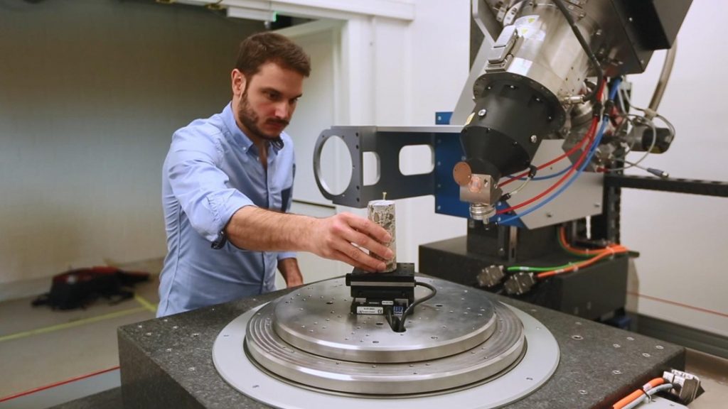 Technician placing cylindrical rock sample of platform below CT scanner © EPFL