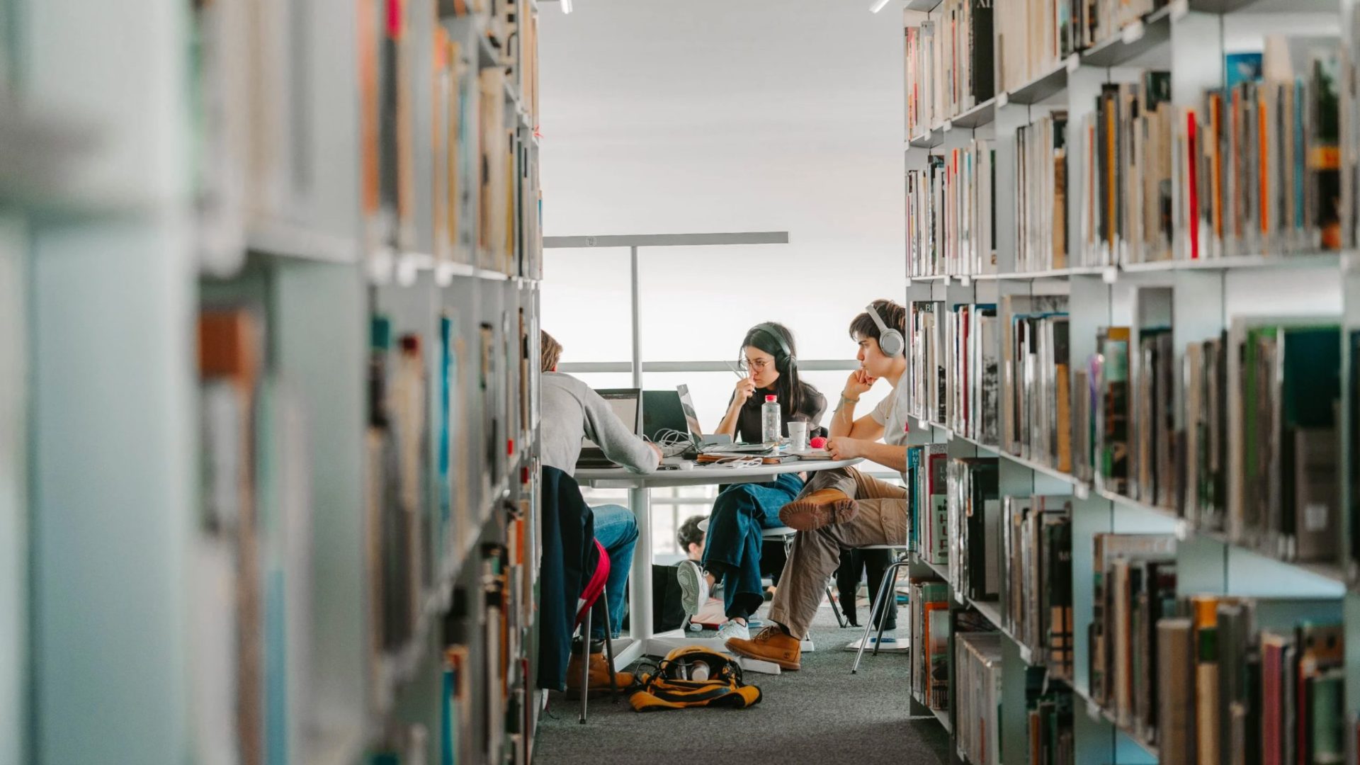 Des étudiantes et étudiants, assis à une table au fond d'une rangée de livres d'une bibliothèque © DR